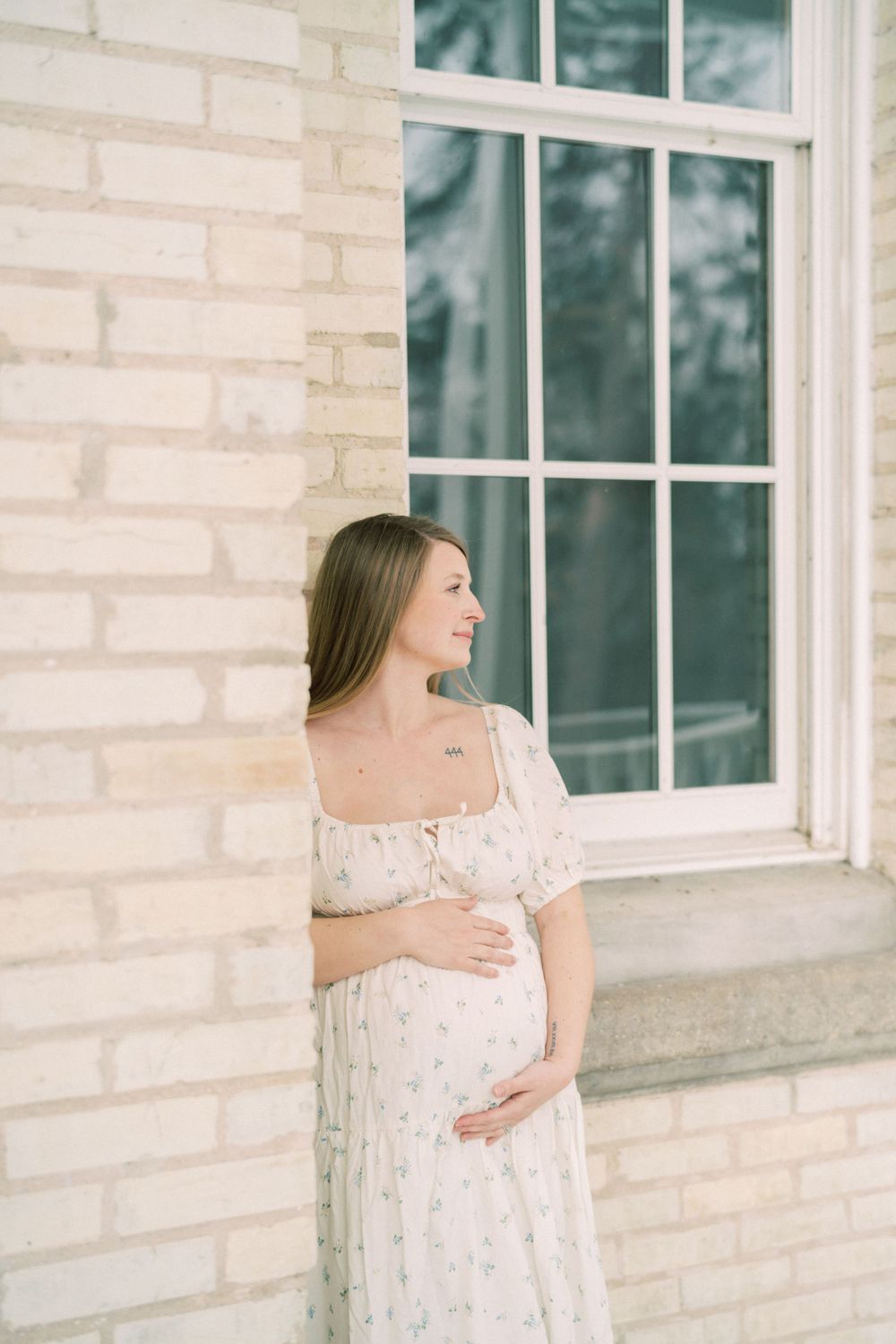 An expectant mother in a white floral dress stands by a window against a light brick wall in soft lighting.