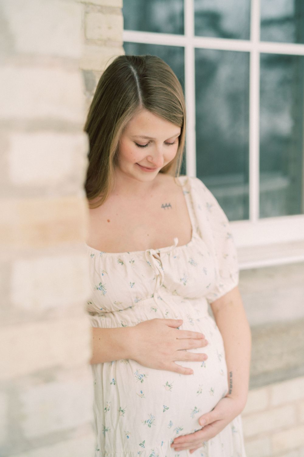 Expectant figure in a white floral dress stands by window in soft natural light during maternity photoshoot.