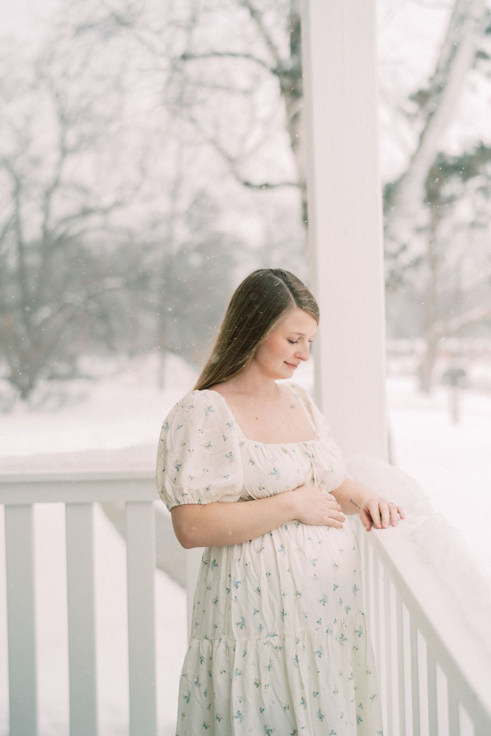 A person in a floral white dress stands on a snowy porch with soft winter lighting in the background.