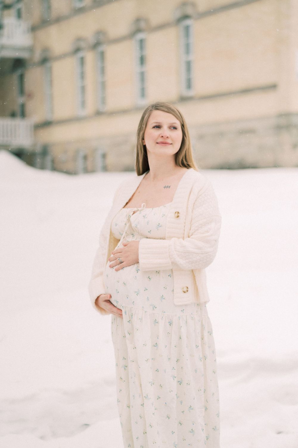 Ethereal winter portrait of someone in a delicate white floral dress and cardigan standing in snow against stone building.