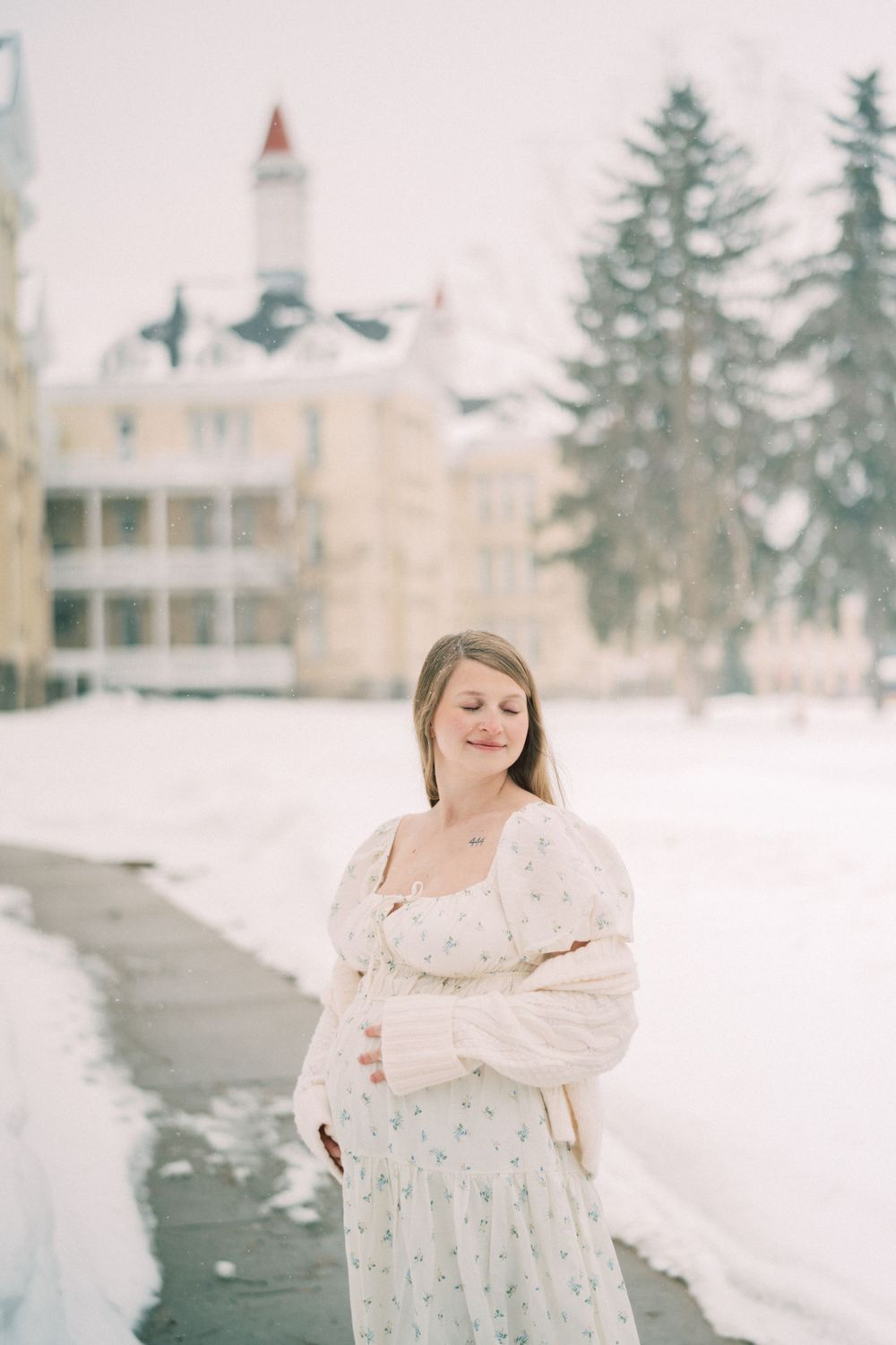 A person wearing a white floral dress stands on a snowy path with a white Victorian building and evergreen trees in background.