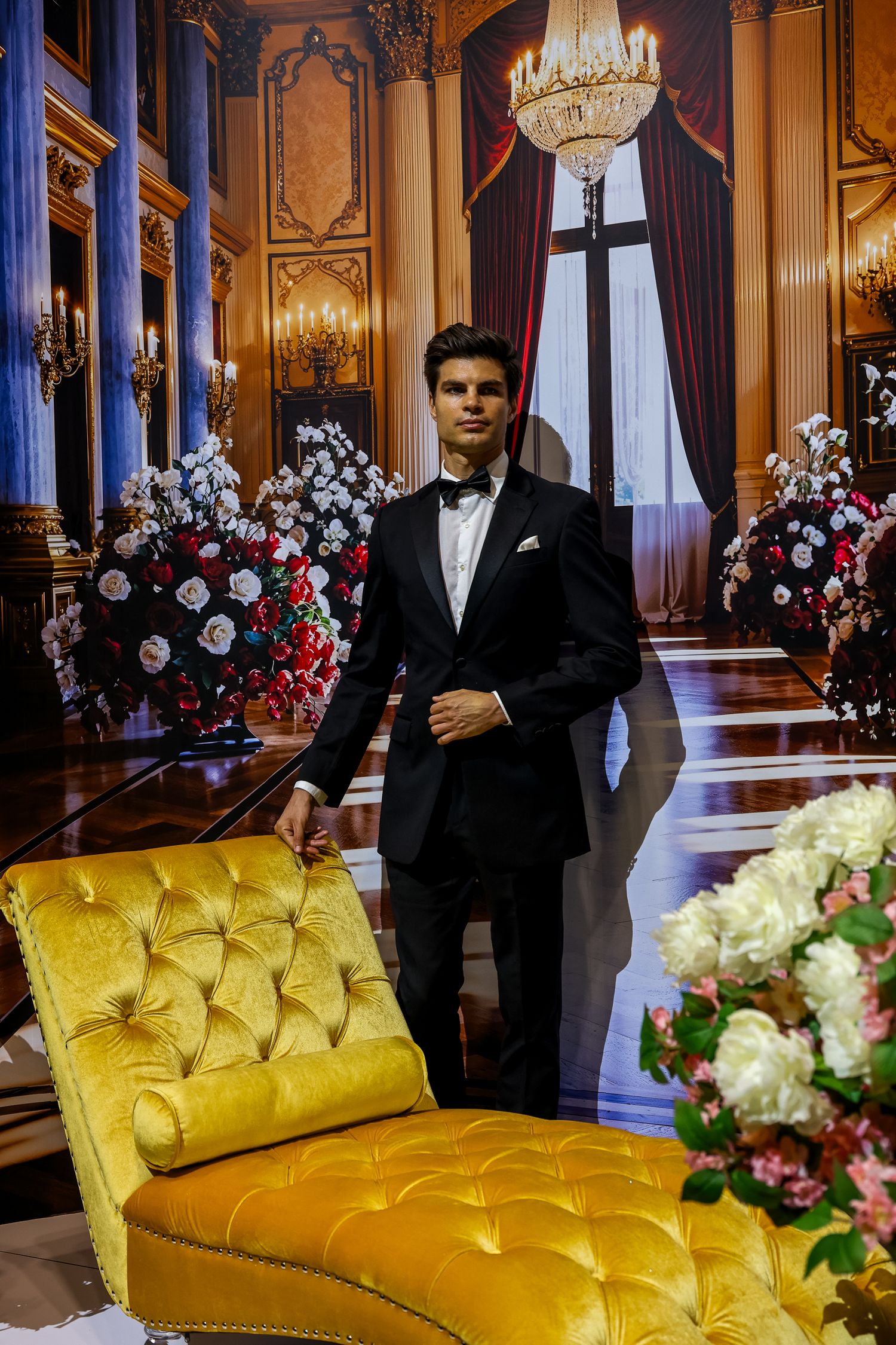 Elegant ballroom setting with gilded walls, crystal chandelier, yellow chaise lounge and floral arrangements in red and white.