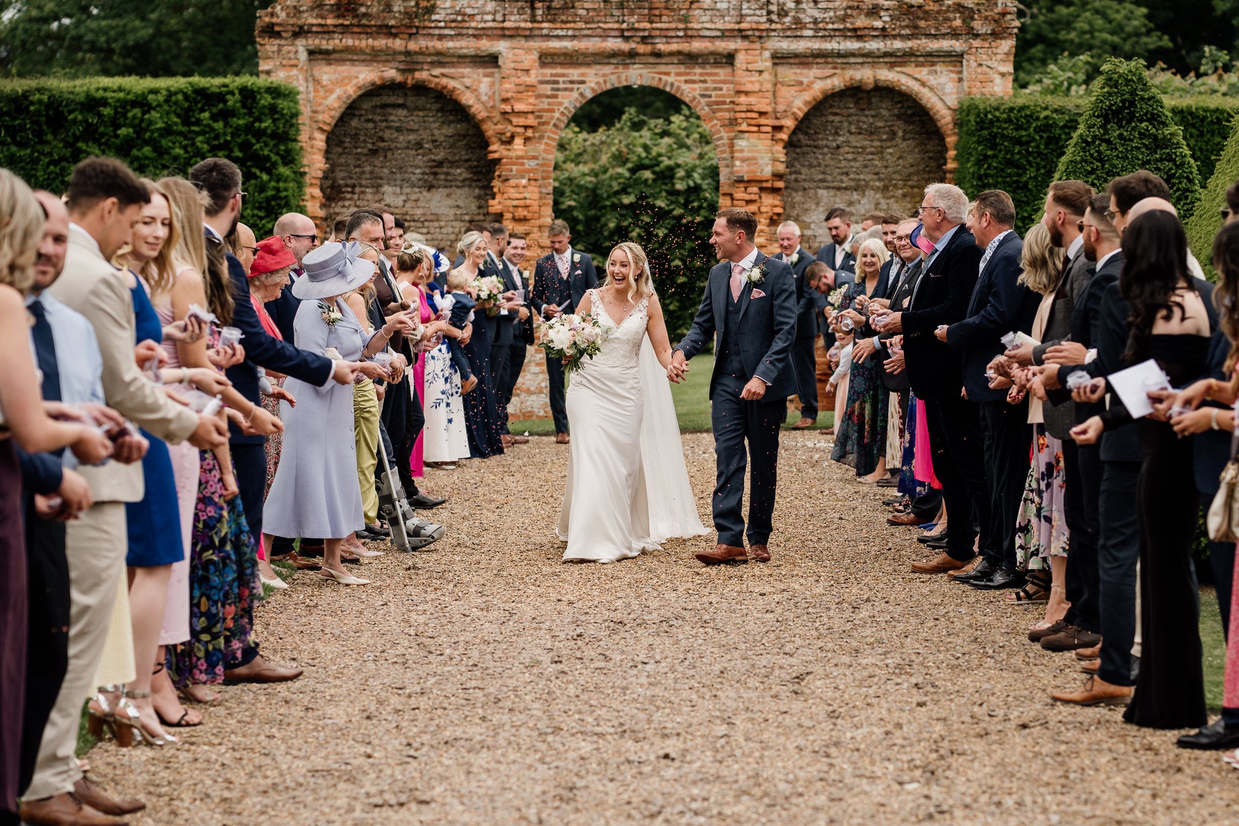 Wedding guests line garden path as couple walks through Oxnead Hall gardens.