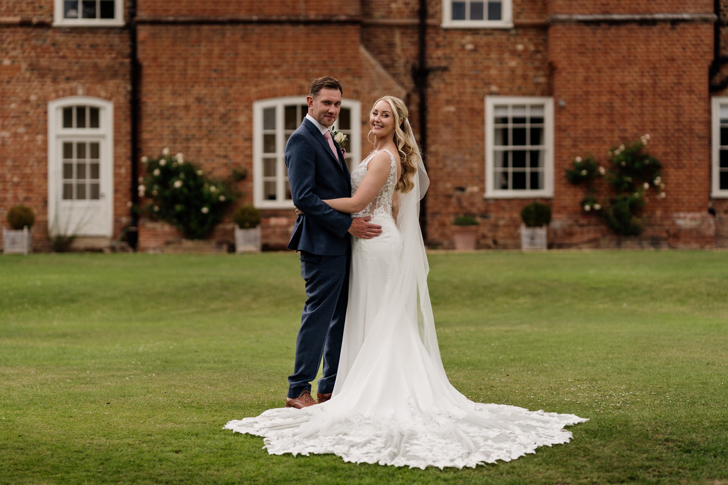 An elegantly dressed married couple stand together on a manicured lawn in front of a historic brick building.