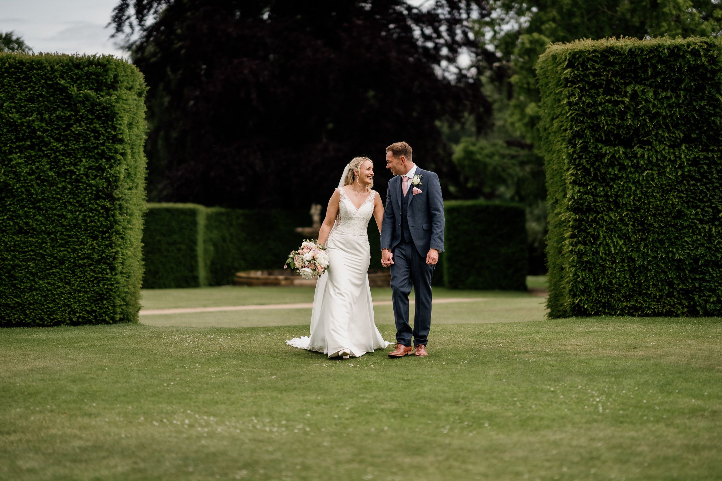A couple in wedding attire walk together on a manicured lawn of Oxnead Hall surrounded by tall green hedges on a sunny day.
