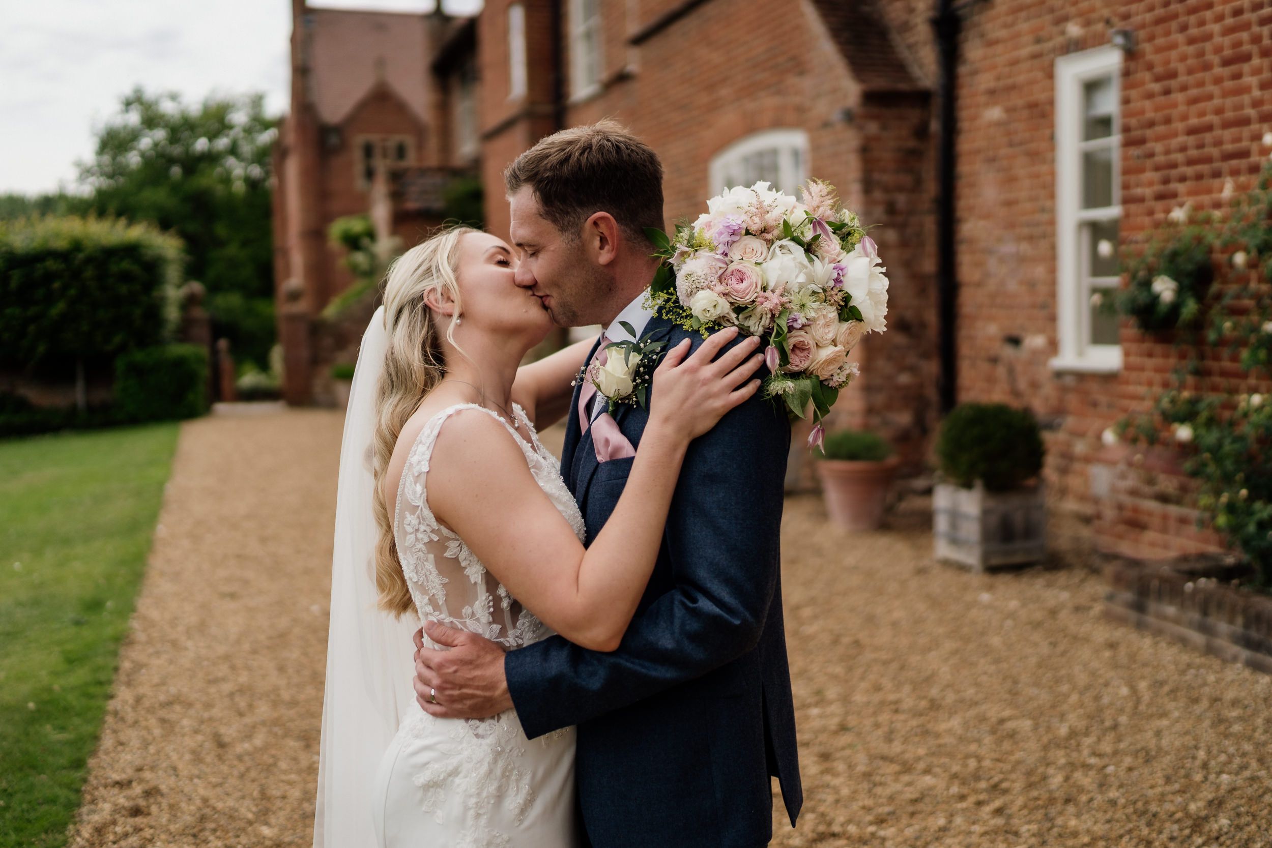 Romantic couple share an intimate moment while standing on gravel path outside brick building with bouquet in hand.