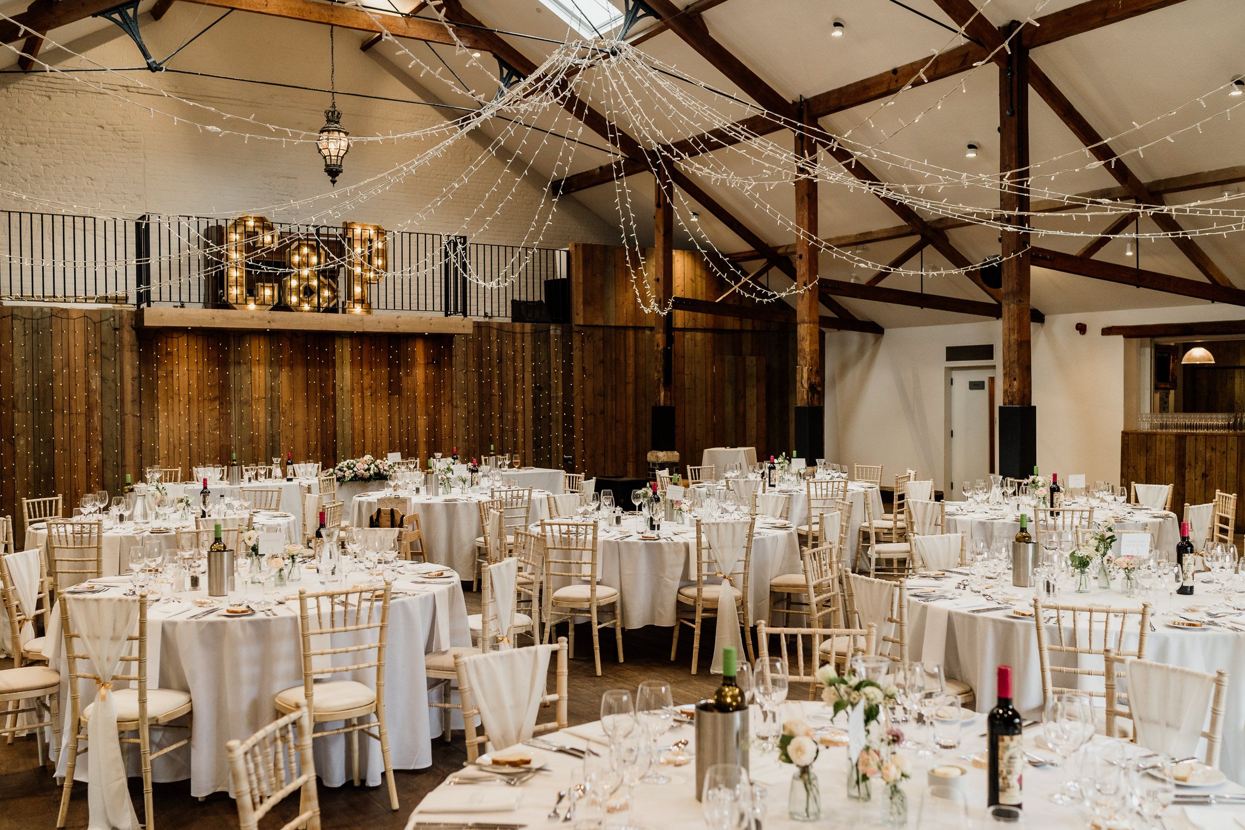 Elegant wedding reception at Oxnead Hall with wooden beams, white tablecloths and chiavari chairs in rustic barn setting.