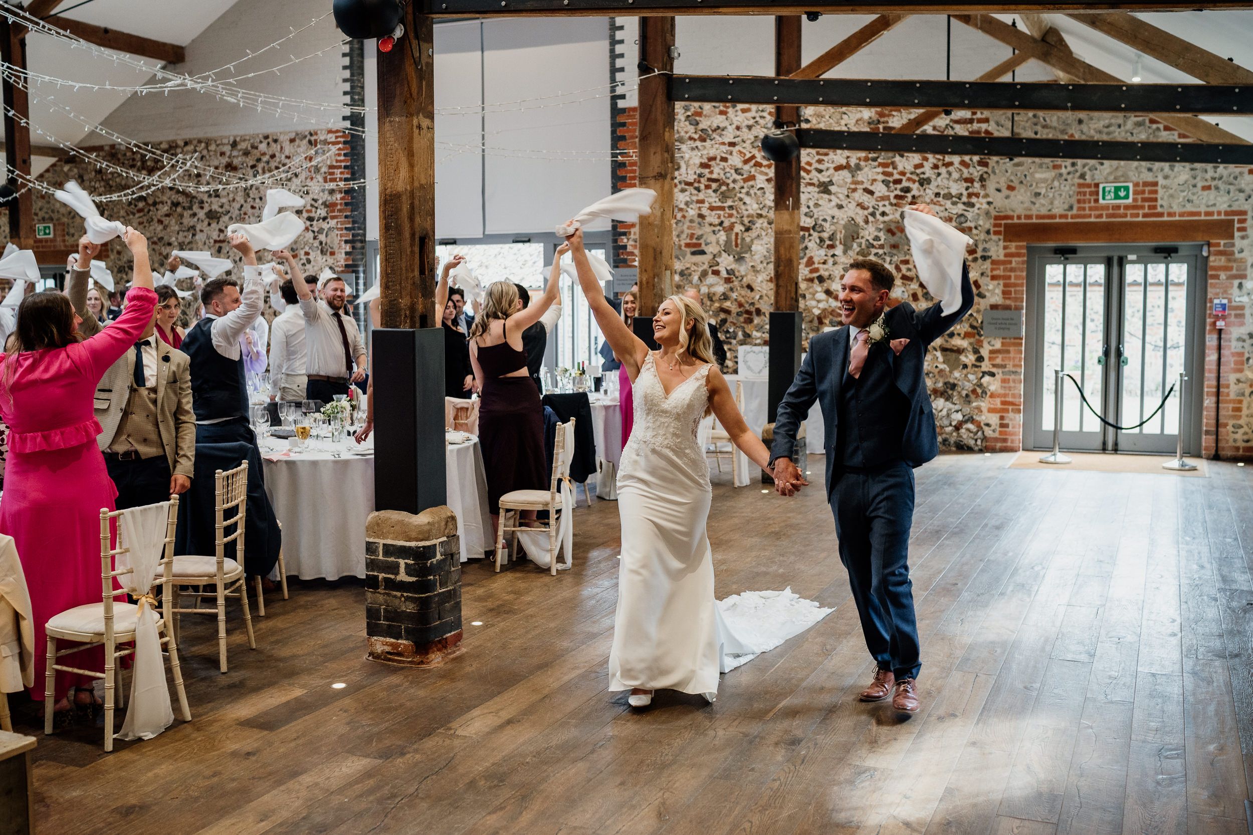 Wedding guests dance and celebrate during a reception in a rustic barn at Oxnead in Norfolk with exposed wooden beams and natural lighting.