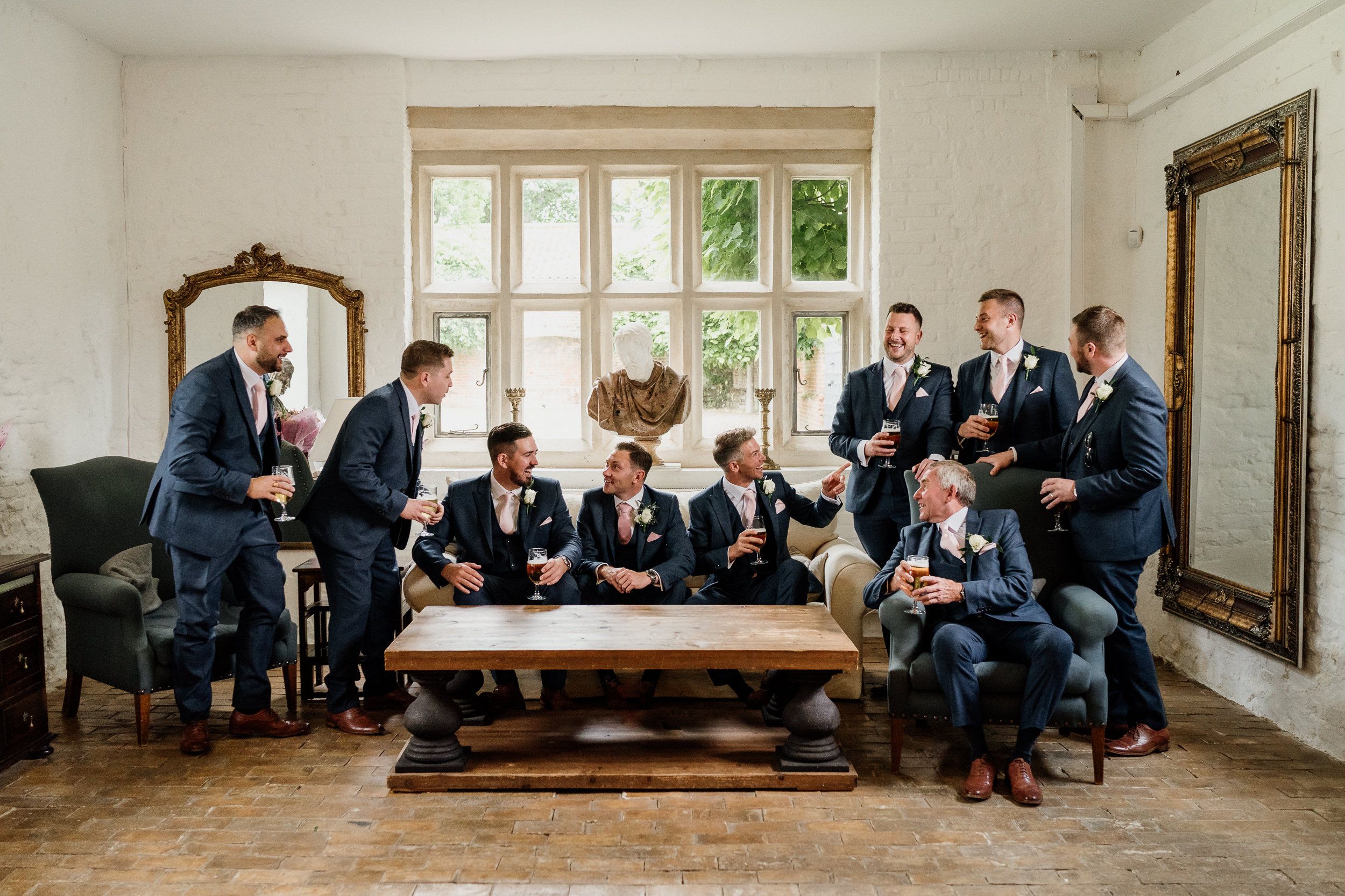 A wedding party group lounges casually in a bright room with large windows and vintage furniture.