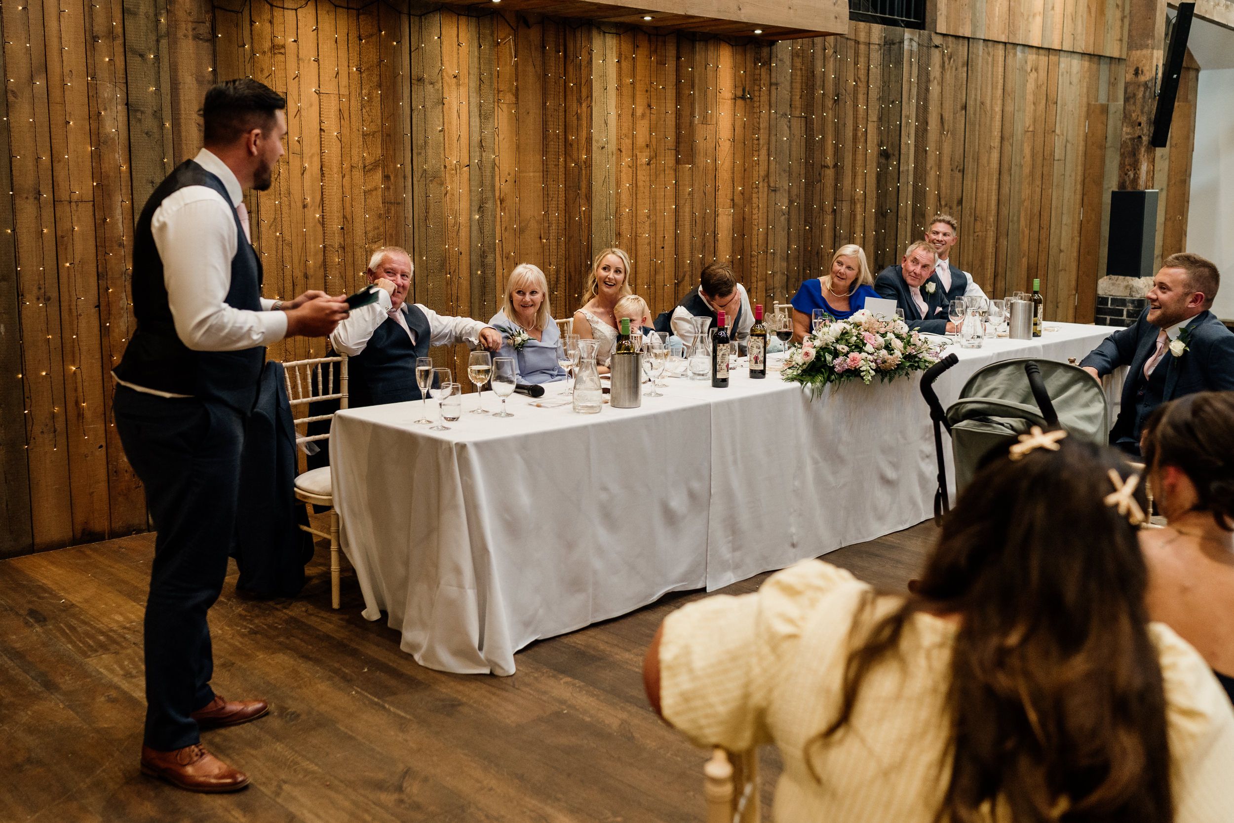 A waiter stands at a long table during a dining event in a rustic wooden venue with guests seated at the table.