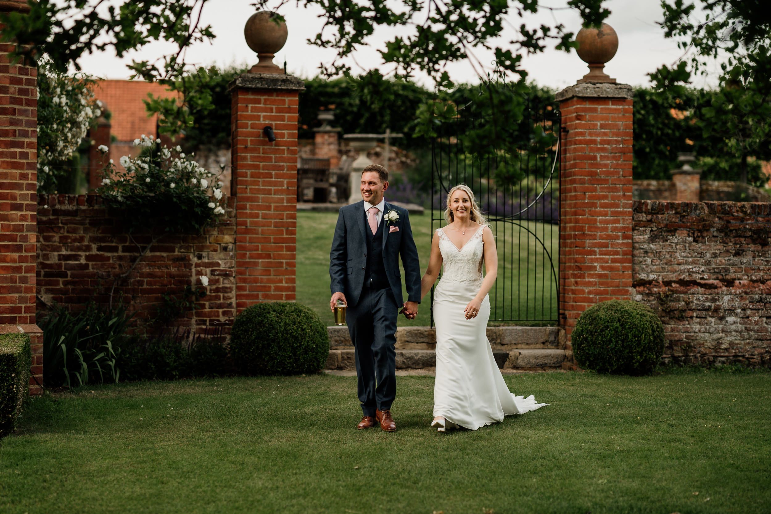 A bride and groom pose together in front of ornate brick gates and a garden wall during their wedding celebration at Oxnead Hall.