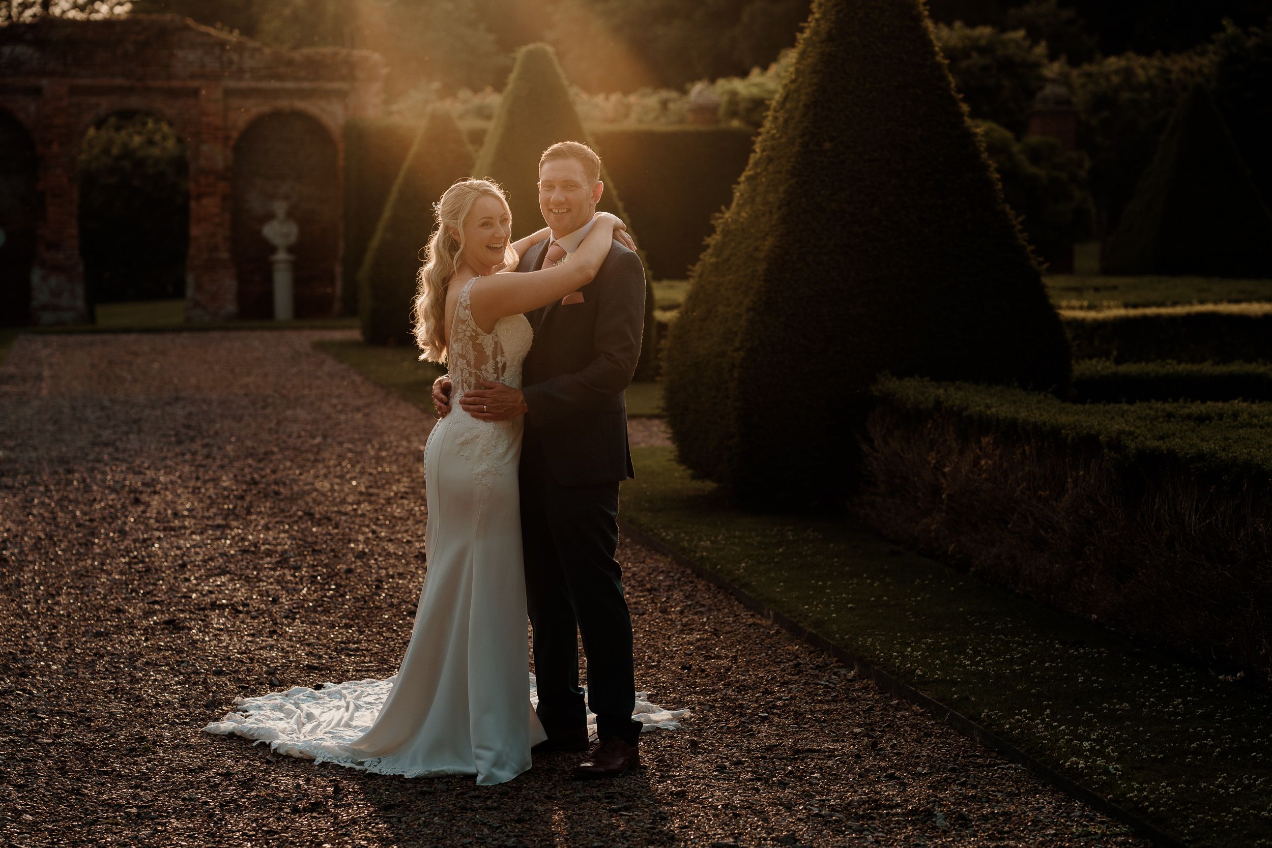 Newlywed couple embraces in romantic sunset moment on garden path with dramatic lighting and historic architecture backdrop.