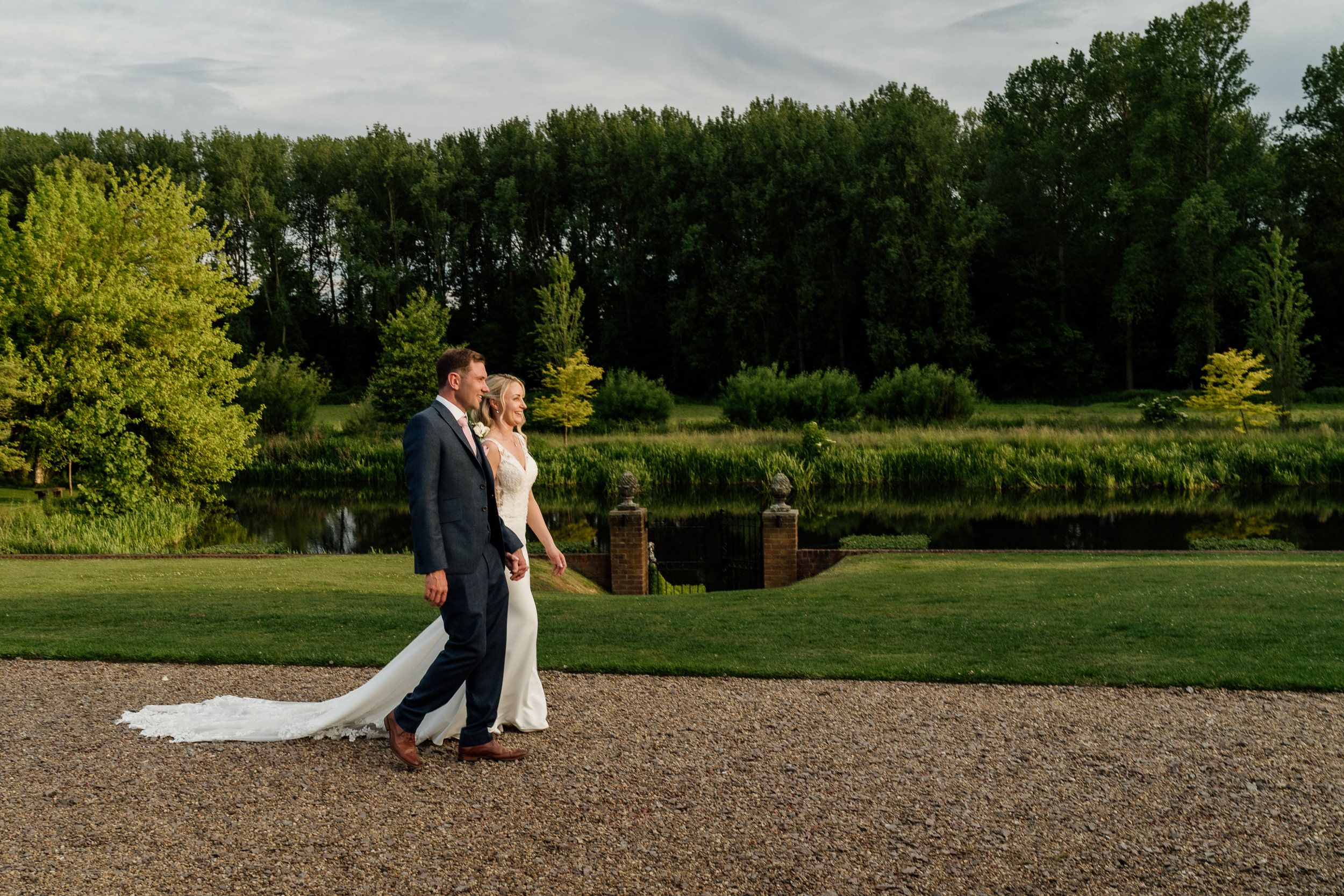 A couple walks in a serene garden setting of Oxnead Hall with lush green trees and natural landscaping on a cloudy afternoon.