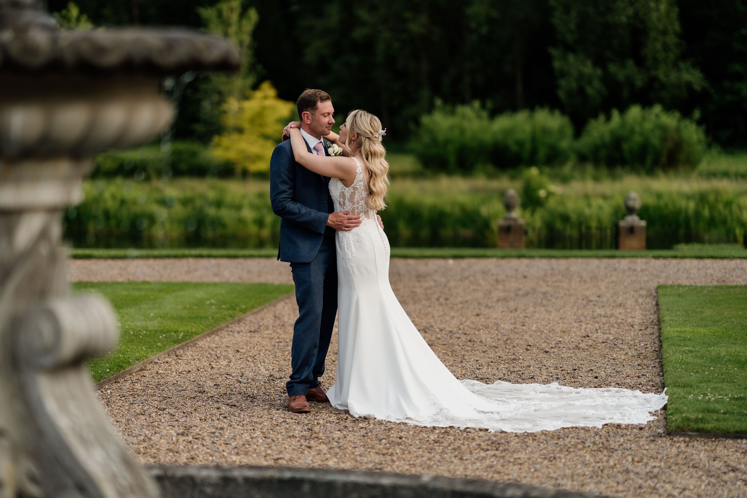 Outdoor garden wedding photo showing romantic couple embracing against lush greenery in formal wedding attire.