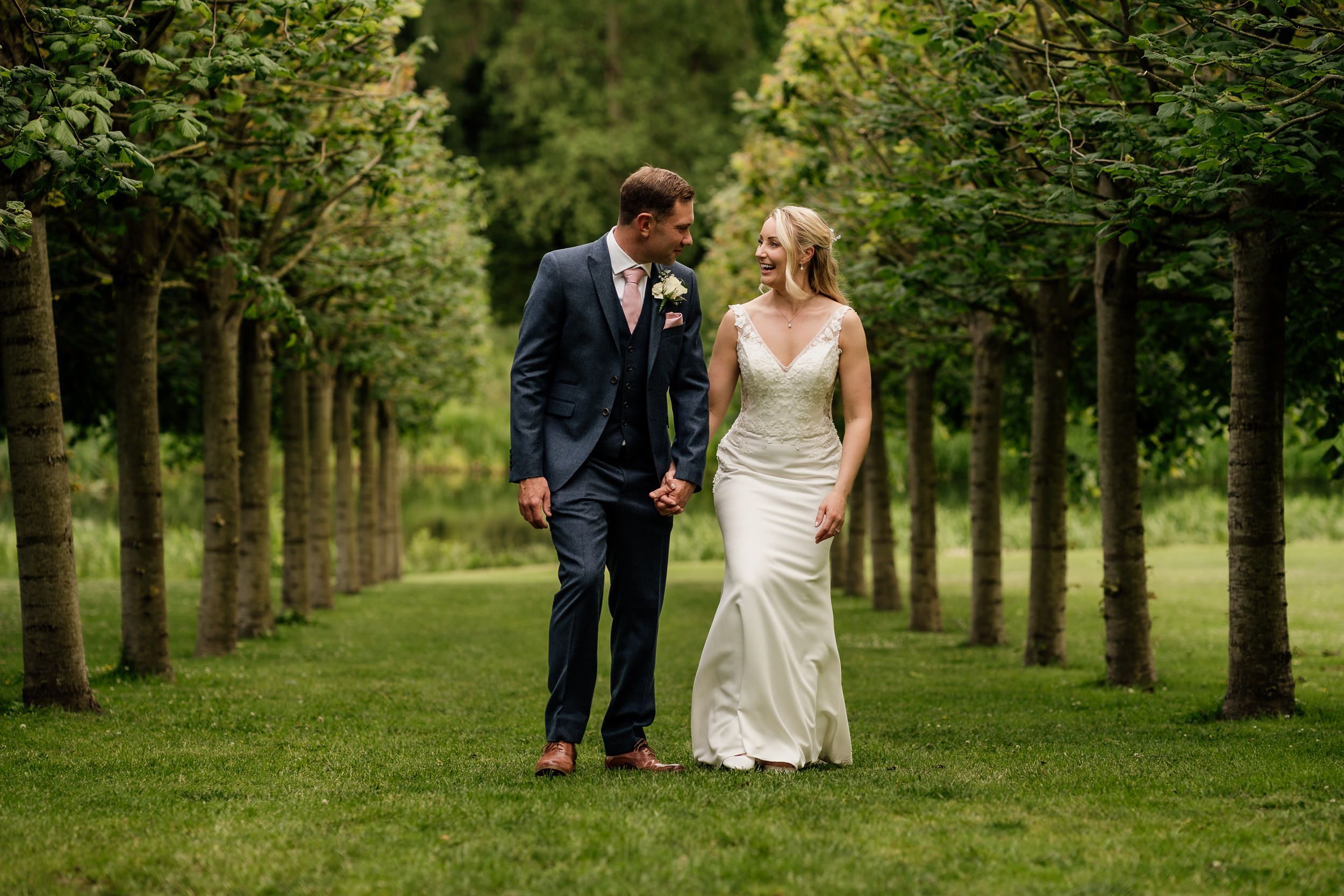 Romantic couple walks hand in hand down a tree-lined pathway with beautiful summer greenery creating a natural tunnel.