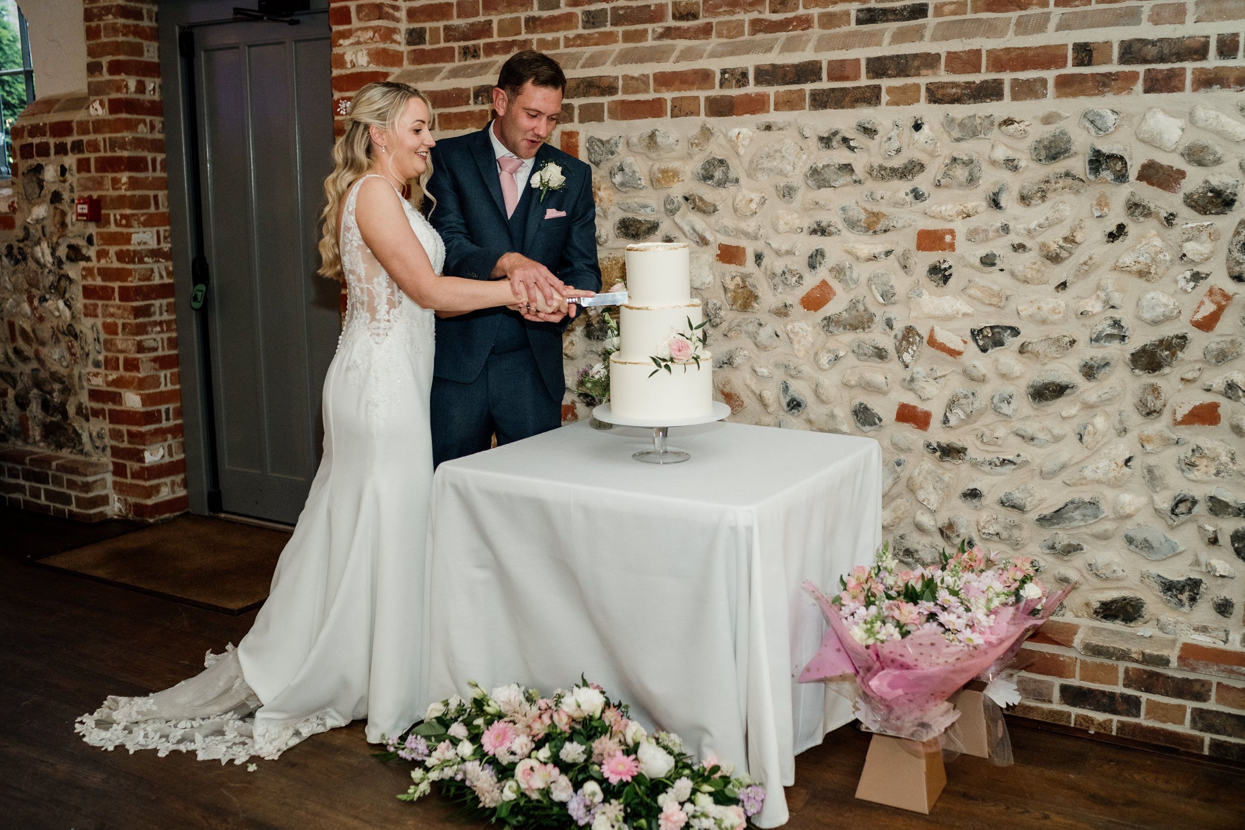Couple cutting their elegant white wedding cake together at rustic brick venue with floral decorations.