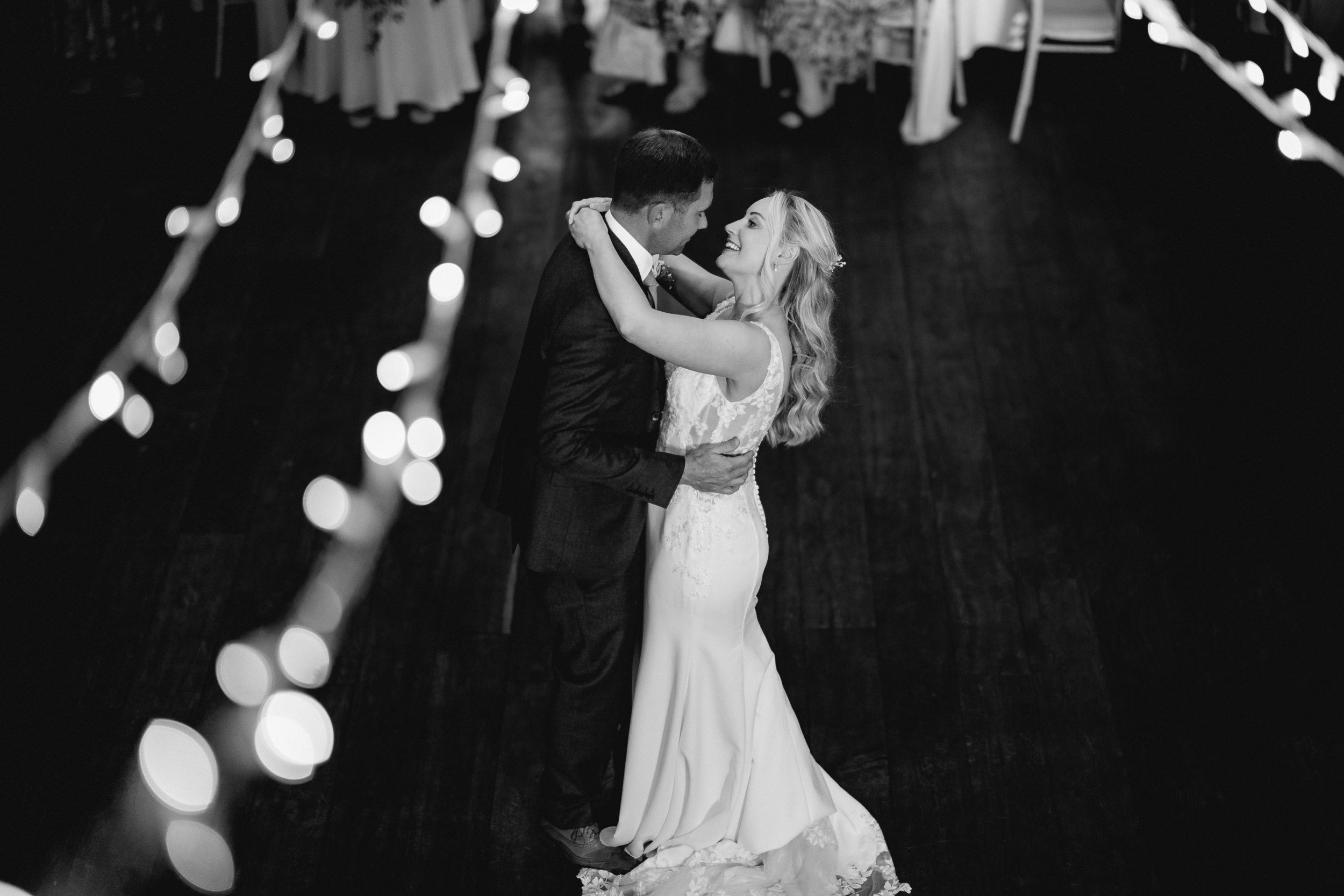 A black and white wedding photo showing a couple dancing at night under string lights.