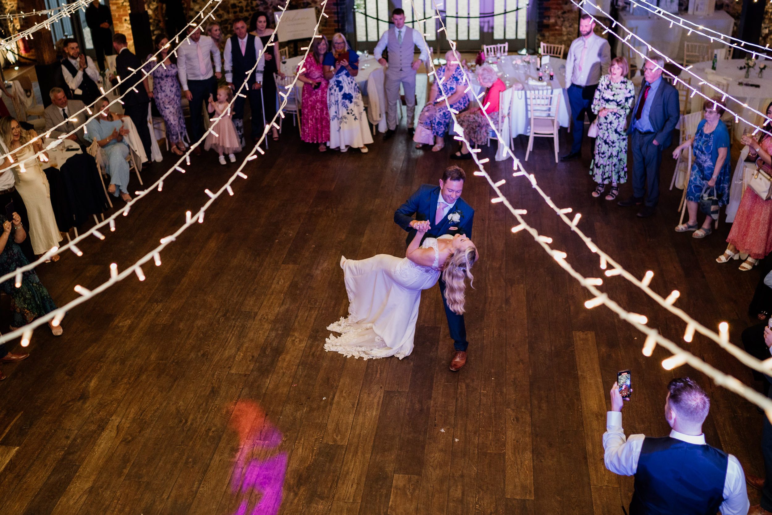 A couple shares their first dance at Oxnead Hall on a wooden floor beneath string lights while wedding guests watch from the perimeter.