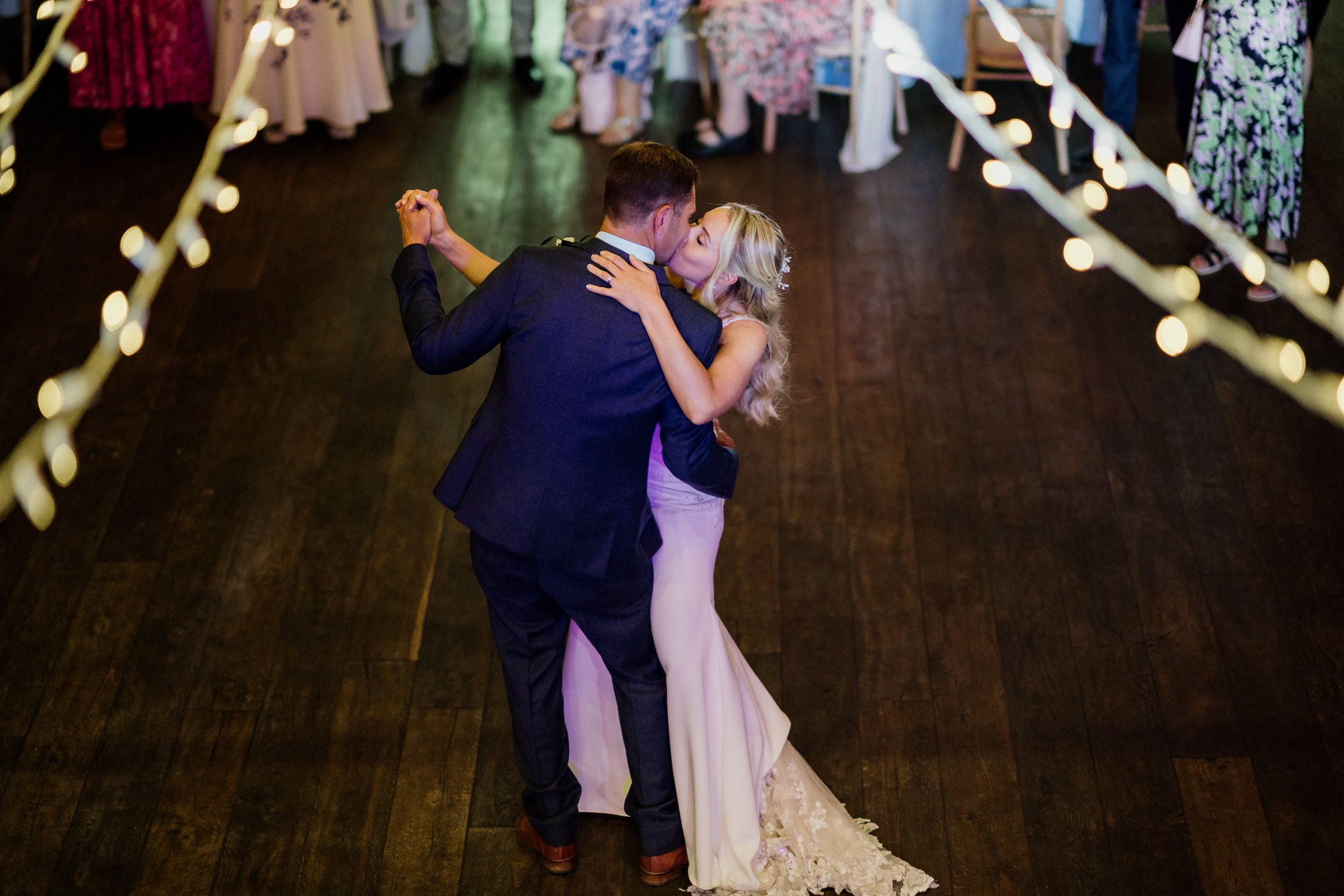 Couple shares an intimate first dance on dark wooden floor beneath string lights at wedding reception venue.