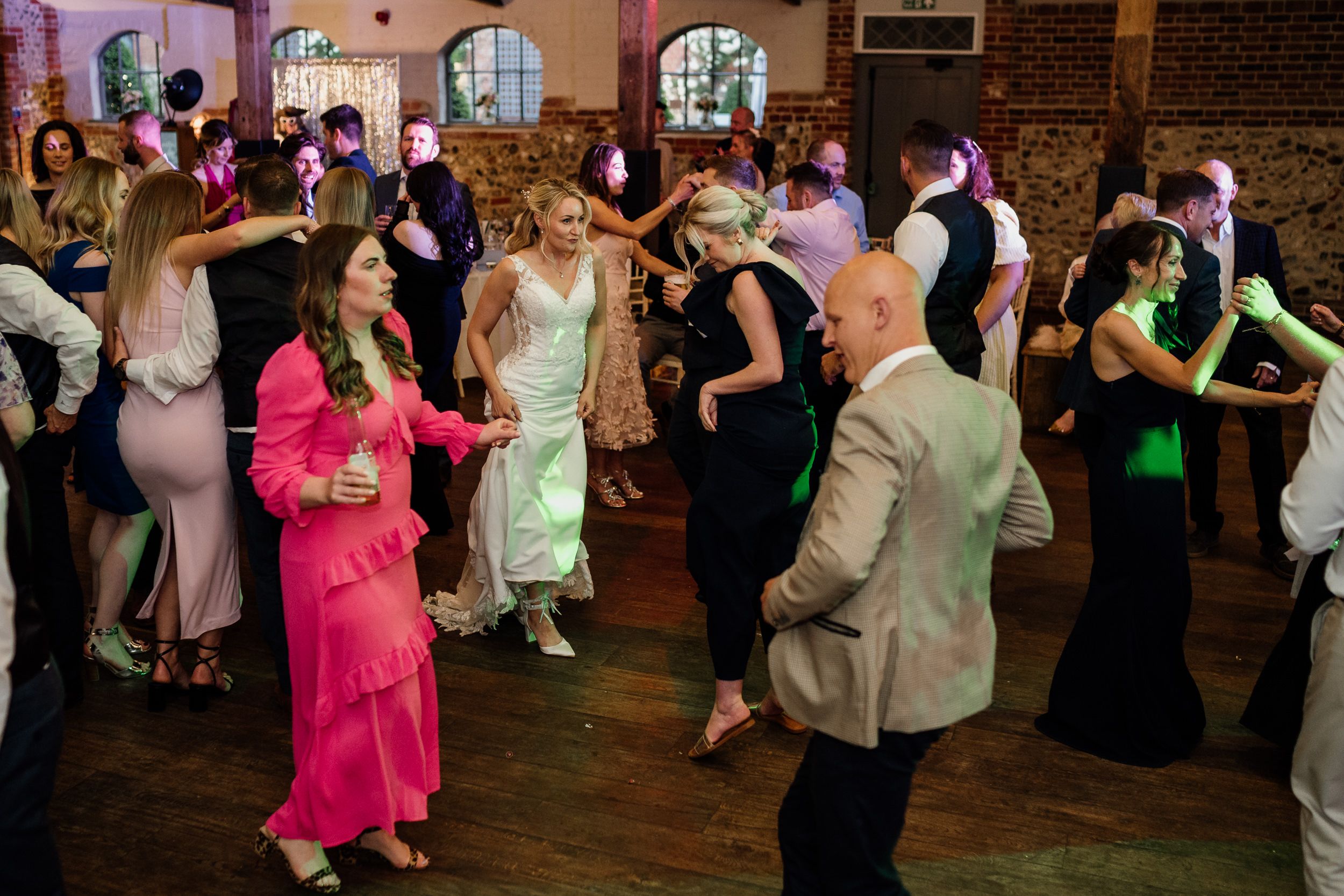 Party guests dance together on a wooden dance floor with colorful lighting in a brick-walled venue at a formal event.