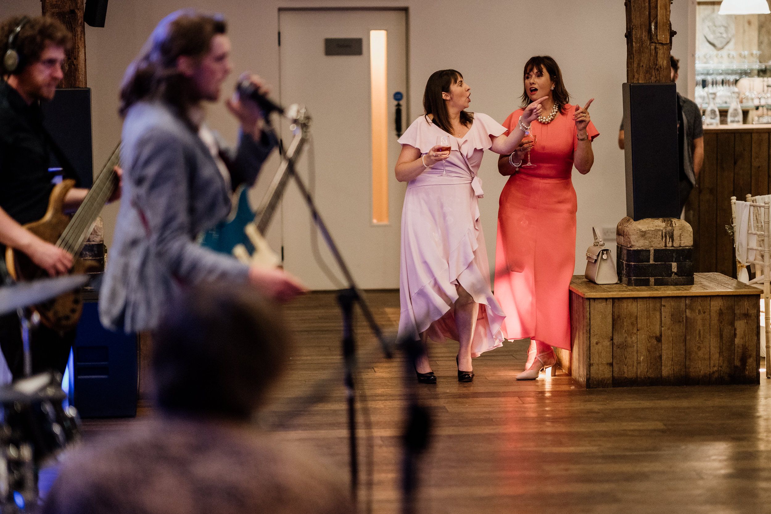 Two performers in pink and coral dresses sing together on stage while musicians play in the background.