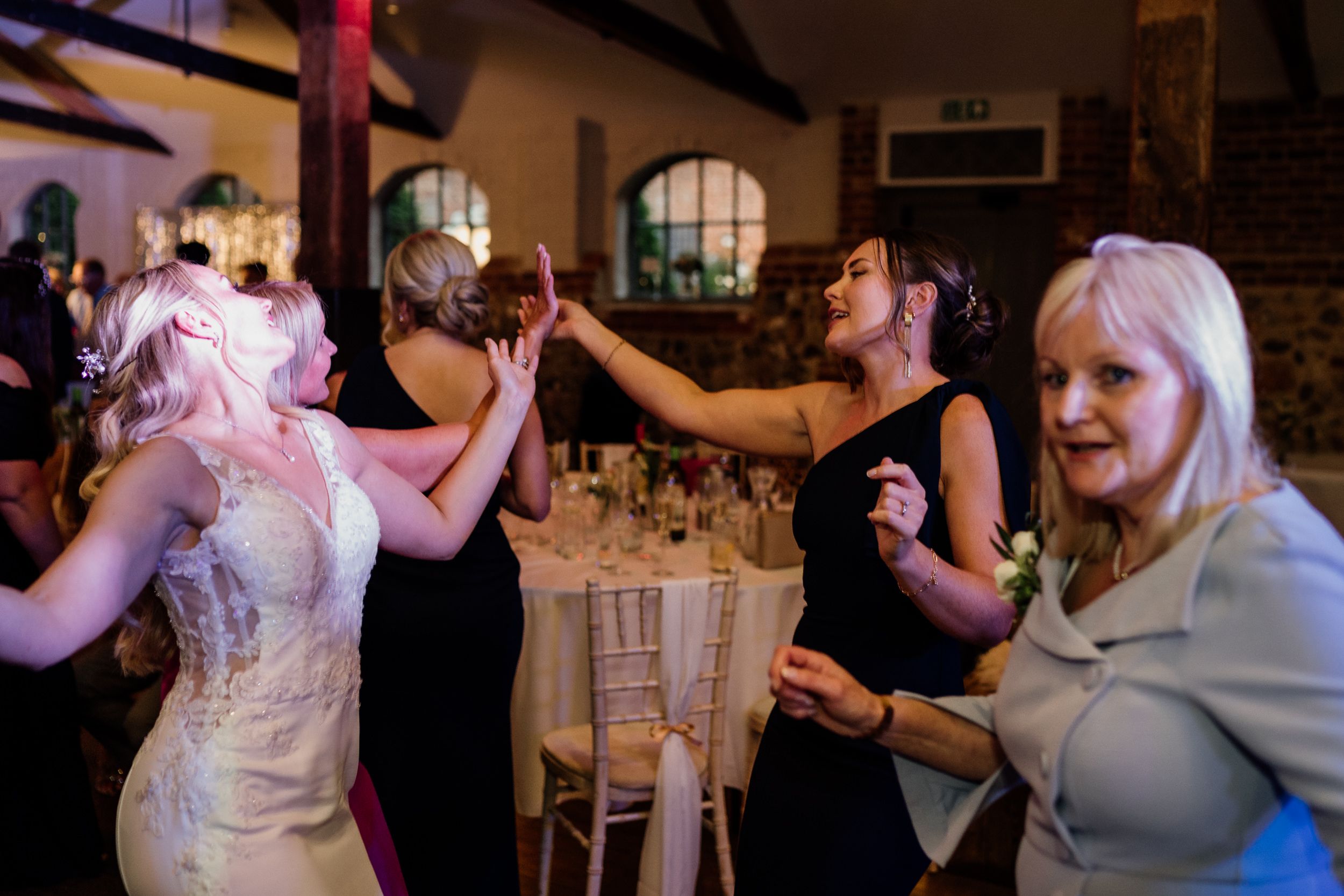 People dancing and enjoying themselves at an indoor wedding reception with brick walls and arched windows.
