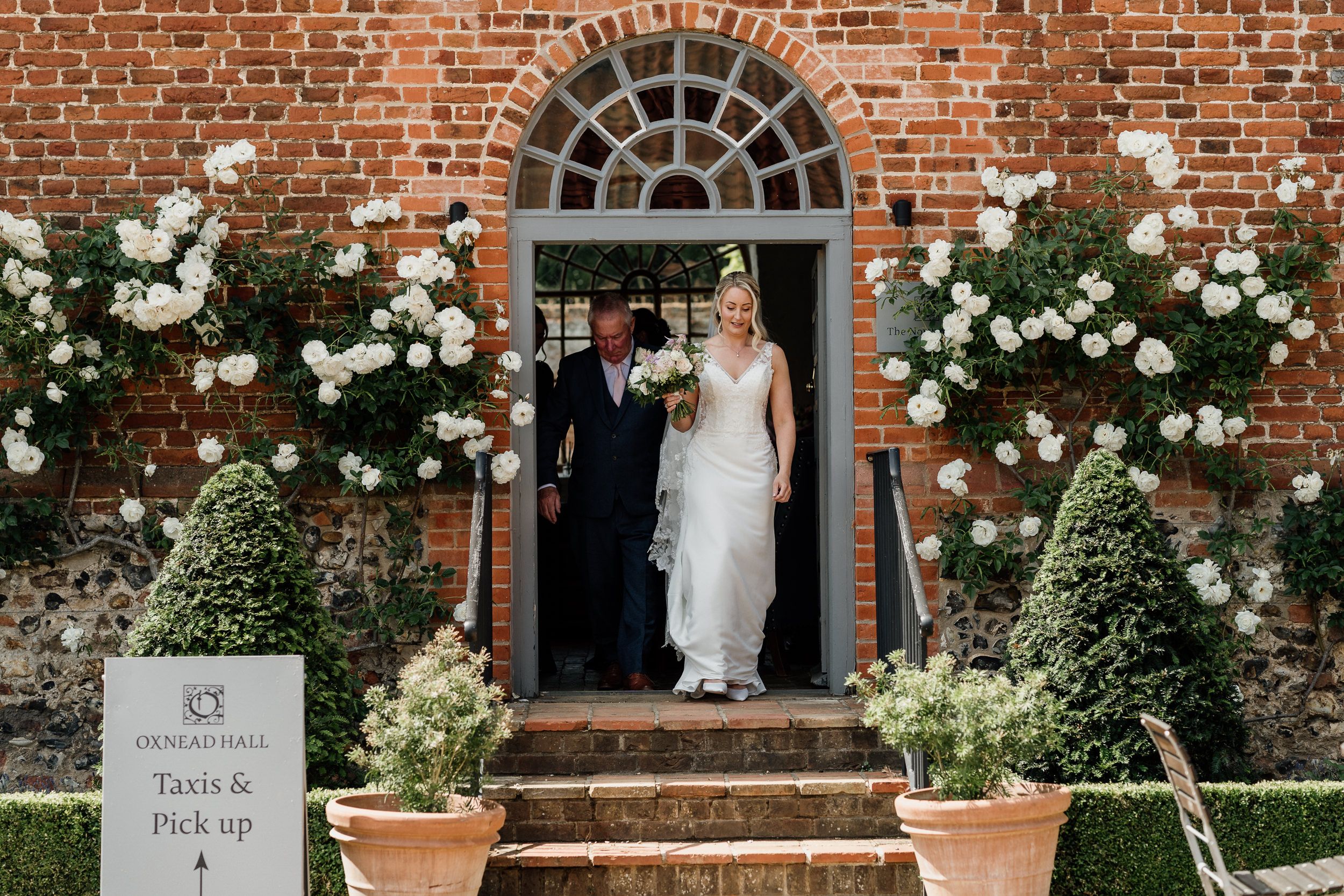 Bride stands in ornate brick doorway adorned with white climbing roses and potted topiaries - Oxnead Hall Wedding.