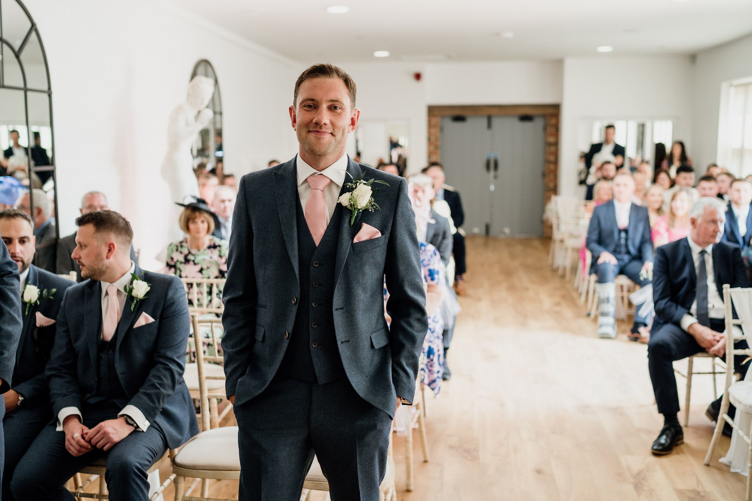 A line of wedding guests in dark suits greet people in a bright, modern venue of Oxnead Hall.