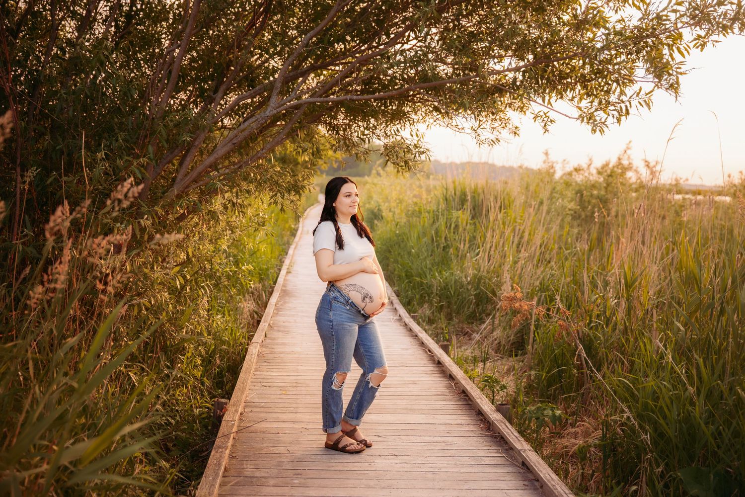 A figure stands on a wooden boardwalk path surrounded by tall grass and trees at golden hour.