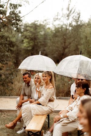 A group of wedding guests sit on wooden benches holding clear umbrellas during an outdoor ceremony.