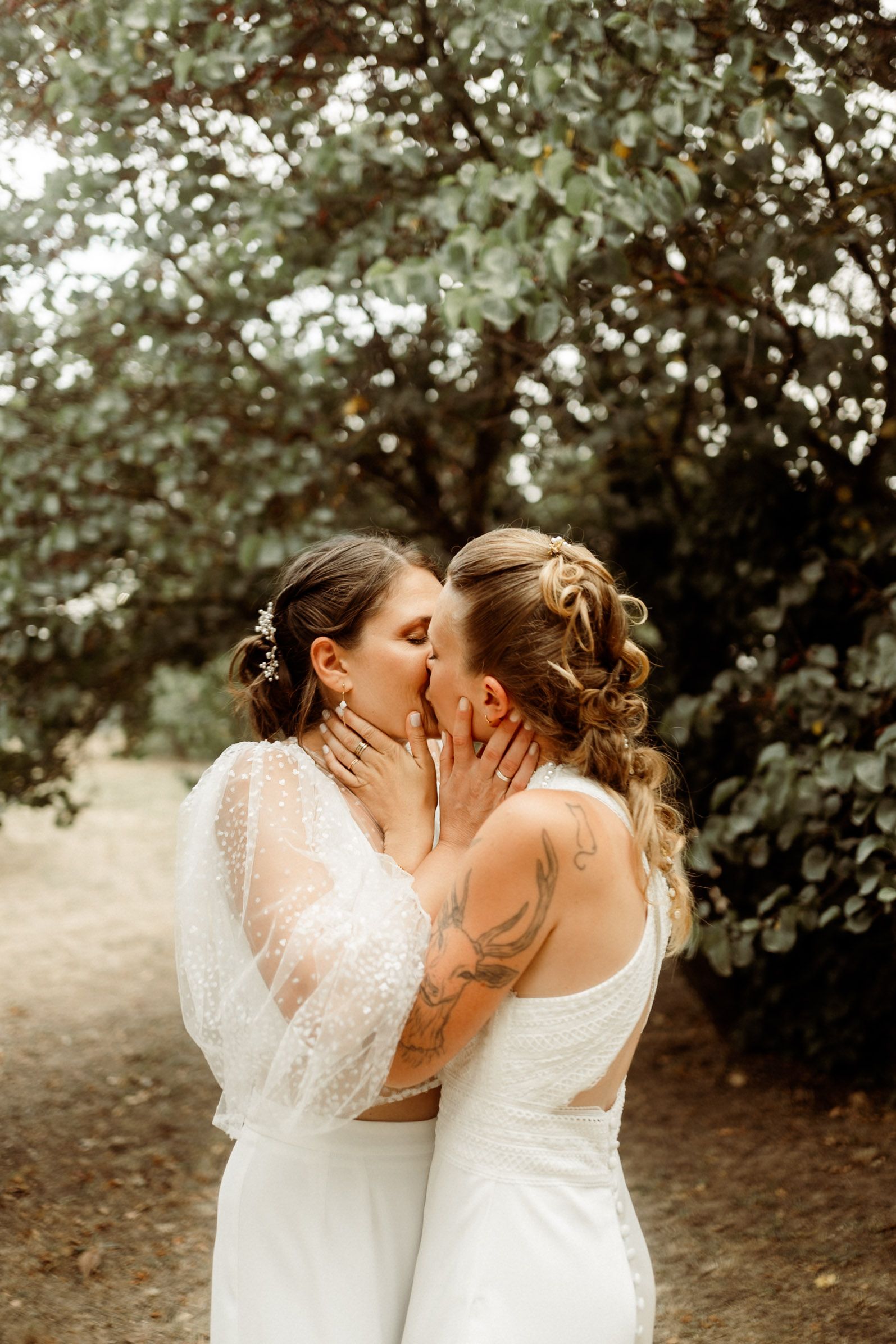 Two people in white wedding dresses share an intimate moment among trees and greenery in a romantic outdoor setting.