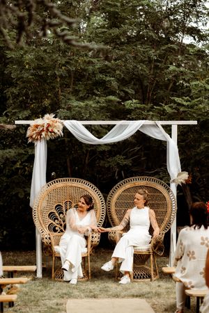 Two people sit in elegant peacock chairs under a white draped arch decorated with dried flowers at an outdoor ceremony.