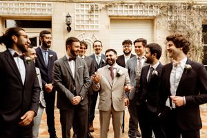 A group of groomsmen in formal attire gather together smiling and laughing outdoors at a wedding event.