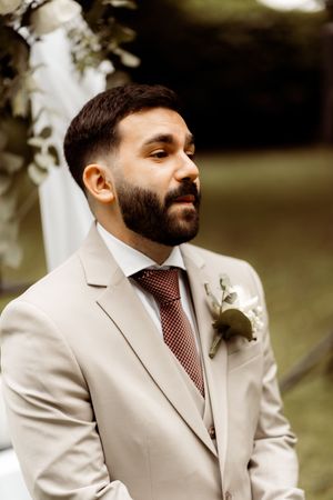 A person in a light beige suit jacket and striped tie stands outdoors with a thoughtful expression.