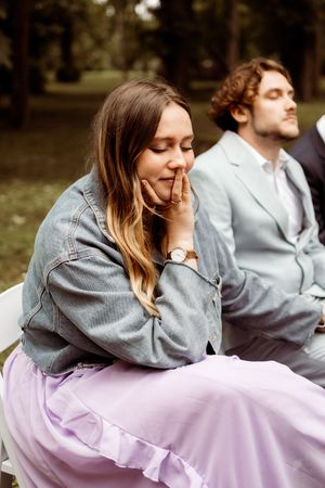 Person in denim jacket and lilac skirt sitting thoughtfully outdoors with another figure in background.