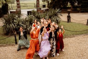 A group of wedding guests in colorful dresses gather excitedly for a celebratory photo outside a historic venue.