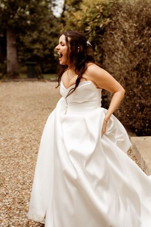 Joyful bride in flowing white wedding dress laughs while standing on gravel path in outdoor garden setting.