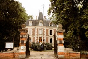 A grand French chateau with ornate architecture and brick pillared entrance gates stands among lush trees and gardens.