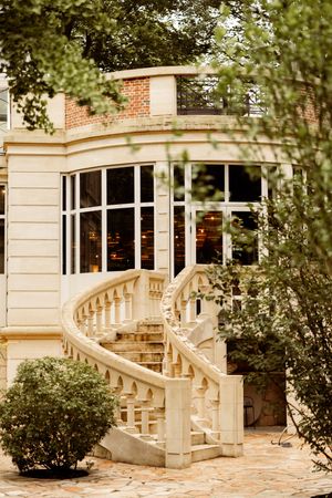 Elegant curved stone staircase leads to a brick building with large bay windows surrounded by lush greenery.