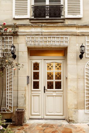 Classic white Parisian building entrance with elegant doors, shutters, and stone facade illuminated by wall lanterns.
