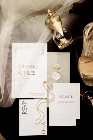 A flat lay of wedding invitation suite with pearl necklace, champagne glass and tulle veil on black background.