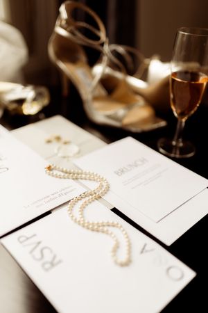 Wedding invitations with pearl necklace and champagne glass in soft focus with gold heels in background.