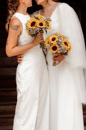 Two brides in white wedding dresses stand close together holding matching sunflower and baby's breath bouquets.