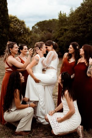 Bridal party in burgundy and white outfits shares candid laughing moment outdoors surrounded by trees.