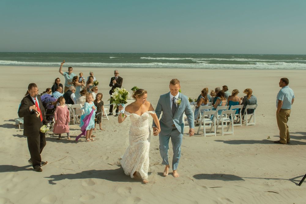 Wedding party celebrating on beach with guests seated on white chairs by ocean.
