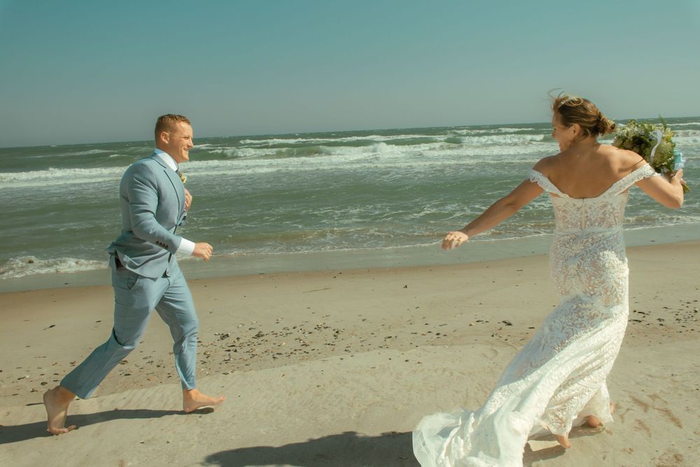 Playful couple running along beach shore in wedding attire.