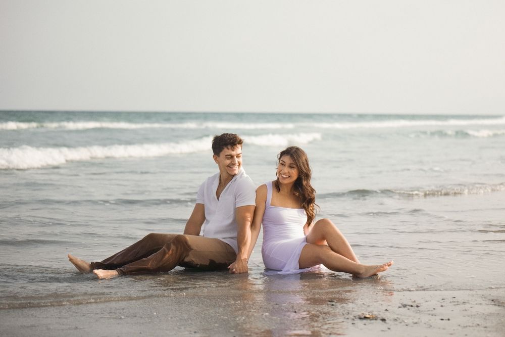 A couple in white outfits sitting together on a sandy beach at sunset.