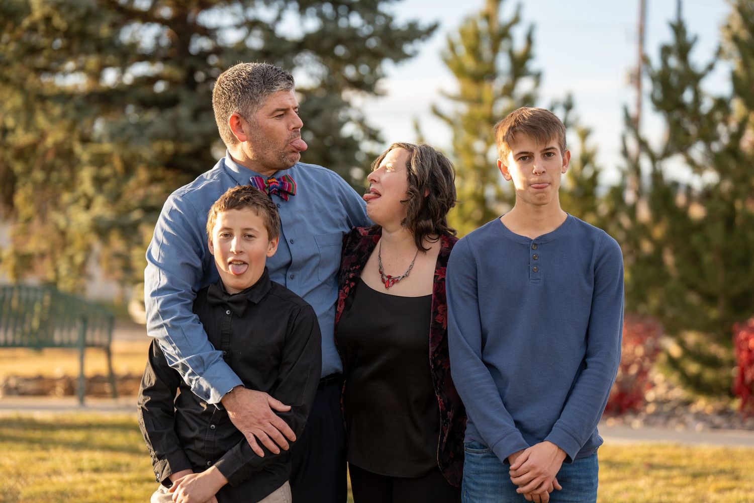 A family of four stands together outdoors during golden hour with evergreen trees in the background.
