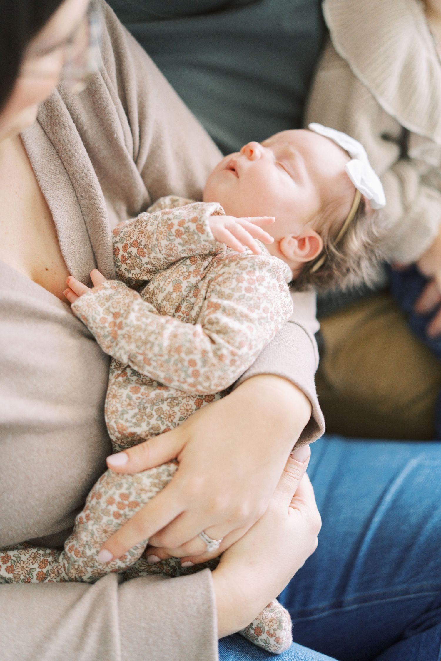 A baby wearing a floral onesie sleeps peacefully in someone's arms on a couch.