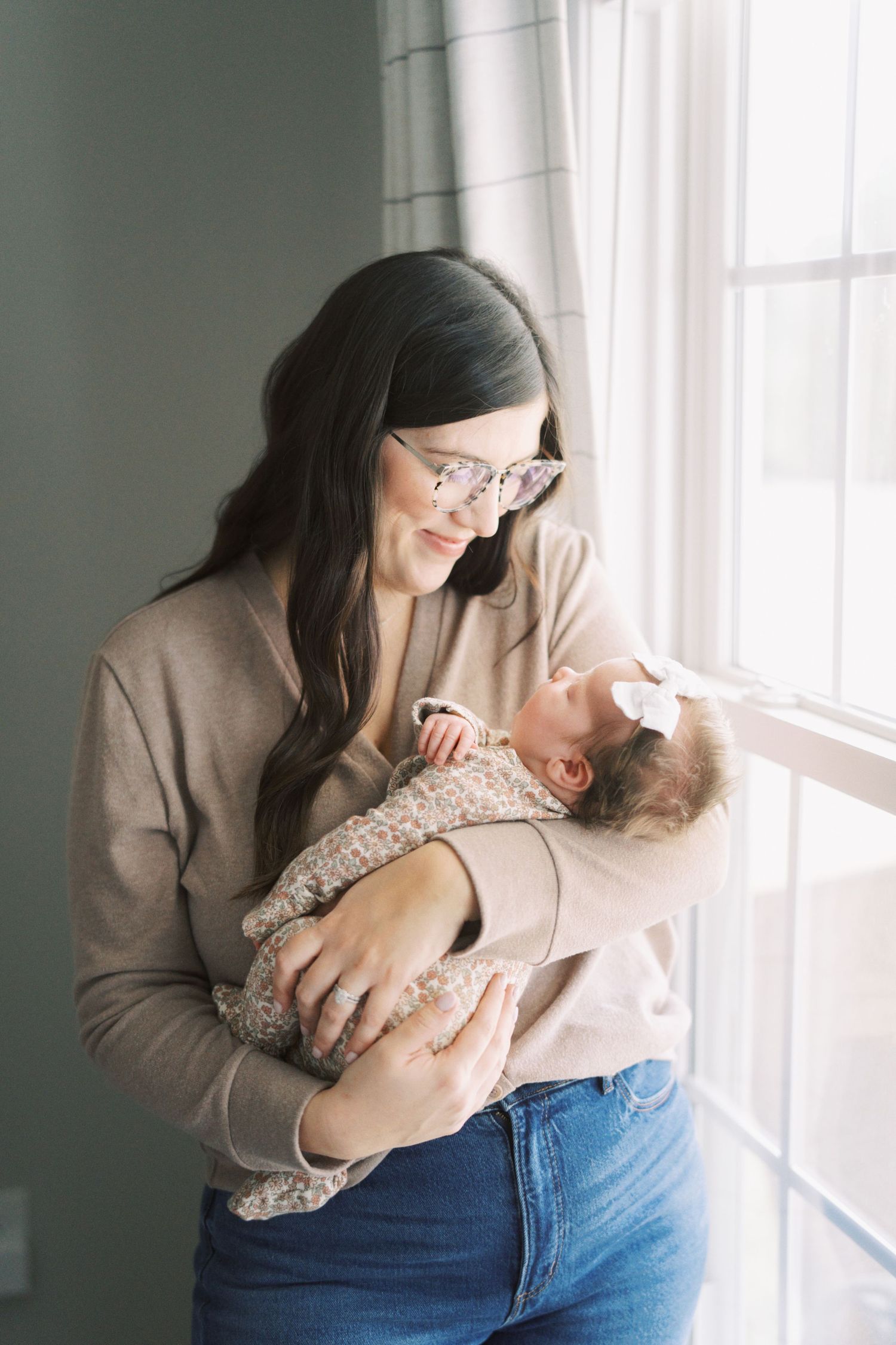 Parent in beige sweater and glasses tenderly holding newborn baby near bright window.